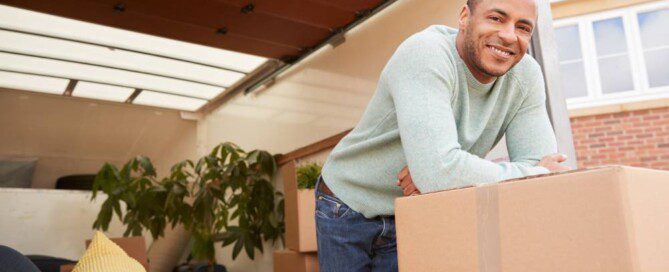 A man leans forward on cardboard boxes in front of a moving truck containing a couch, houseplant, and other cardboard boxes.