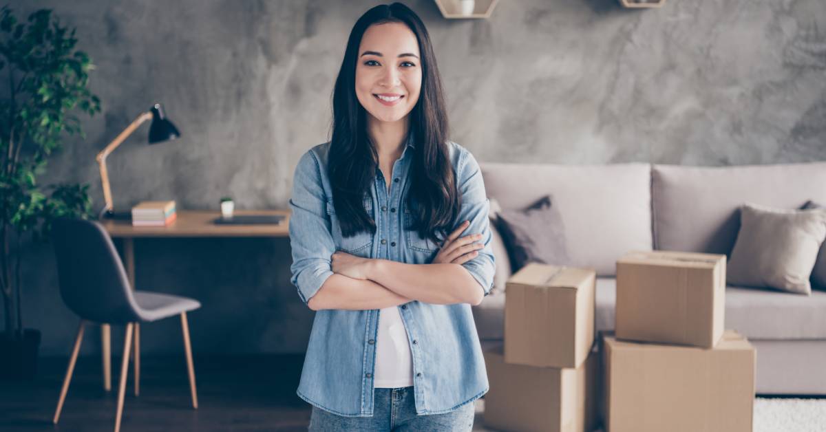 A young Asian woman stands with her arms crossed, smiling in front of a work desk, a couch, and sealed cardboard boxes.