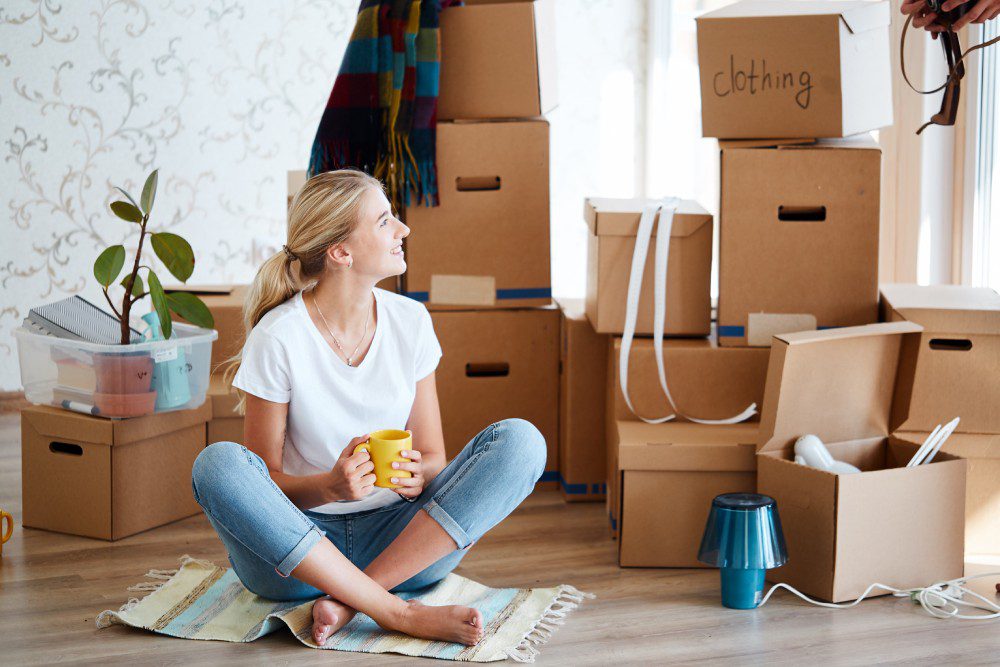 A young woman sitting in her new apartment surrounded by packed cardboard boxes, holding a coffee mug and smiling.