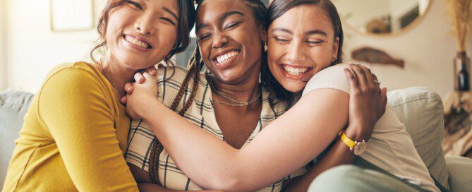 Three young women sit on a couch in a living room and wrap their arms around each other while smiling.