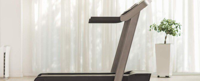 A treadmill sits on an off-white rug with matching flooring, white walls, sheer curtains, and a planter holding a tree.