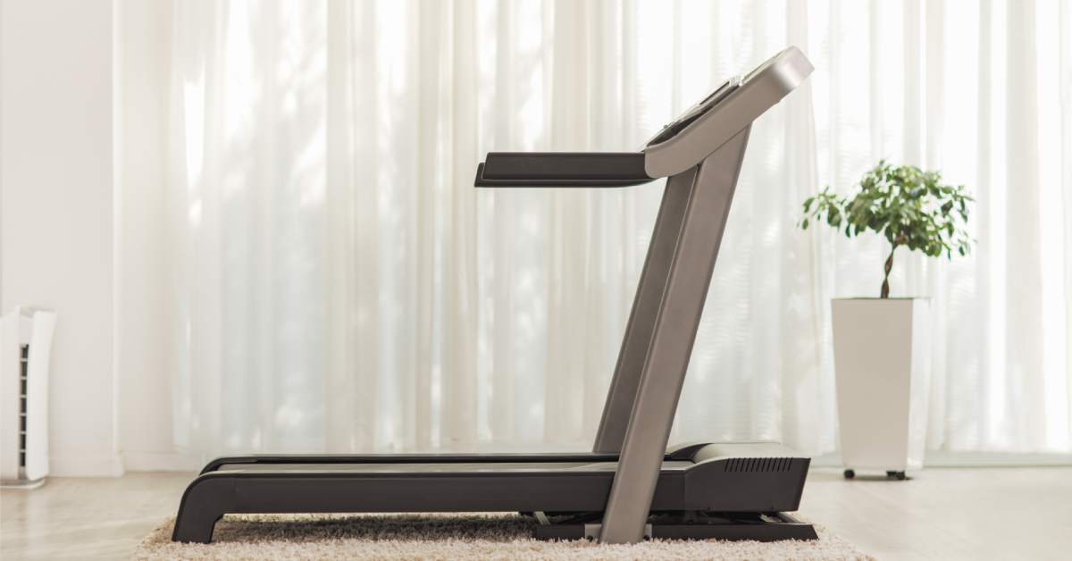 A treadmill sits on an off-white rug with matching flooring, white walls, sheer curtains, and a planter holding a tree.