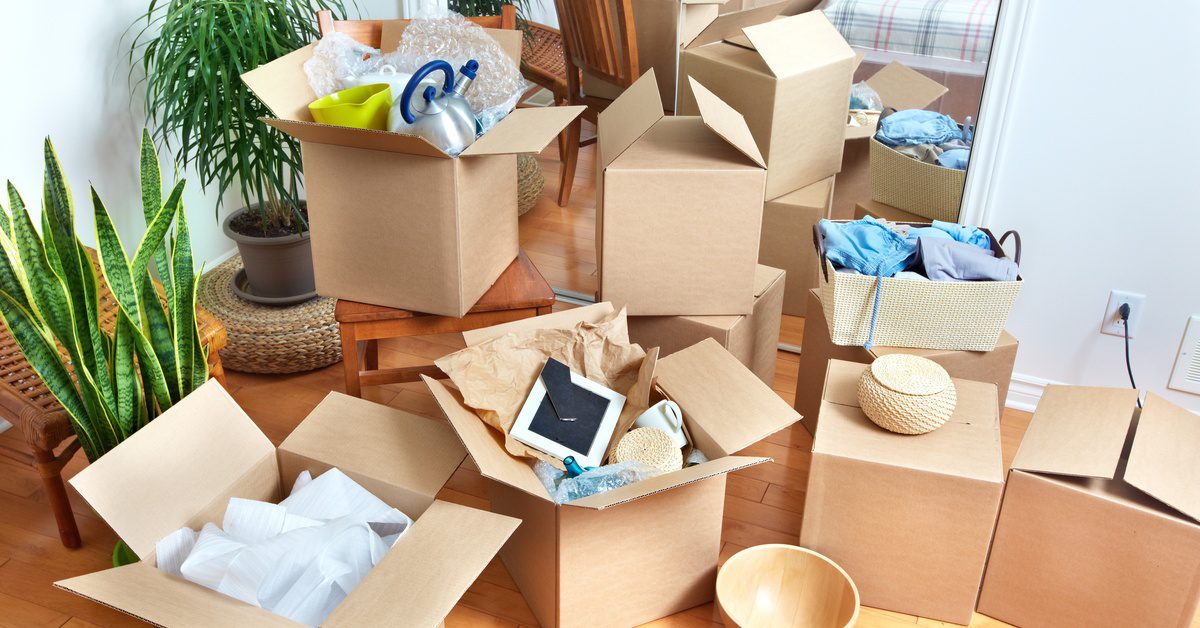 A brightly lit room is full of moving boxes. Some of the boxes are closed while others are overflowing with stuff.
