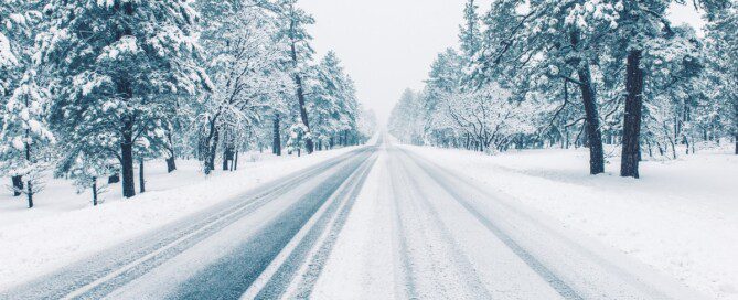 A long, empty road is covered with snow and has tire tracks from cars driving. There are snow covered trees along the side of the road.