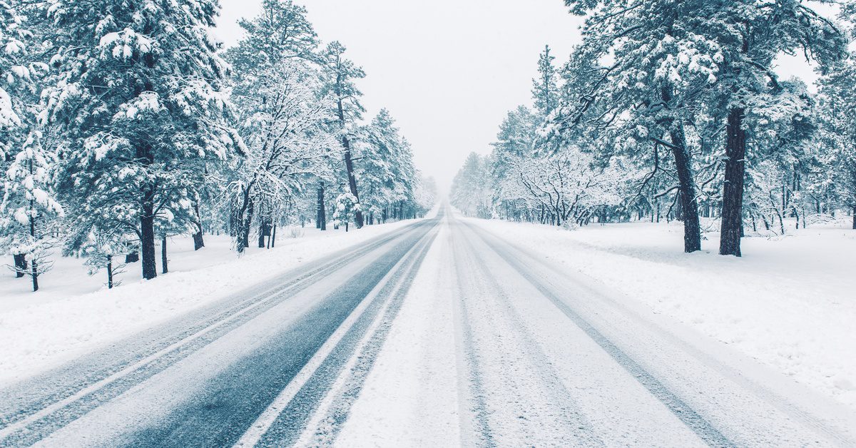 A long, empty road is covered with snow and has tire tracks from cars driving. There are snow covered trees along the side of the road.