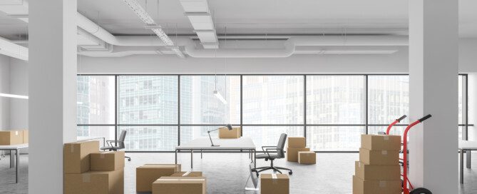 Multiple stacks of cardboard boxes on the gray floor of an open office space with white tables, chairs, walls, and columns.