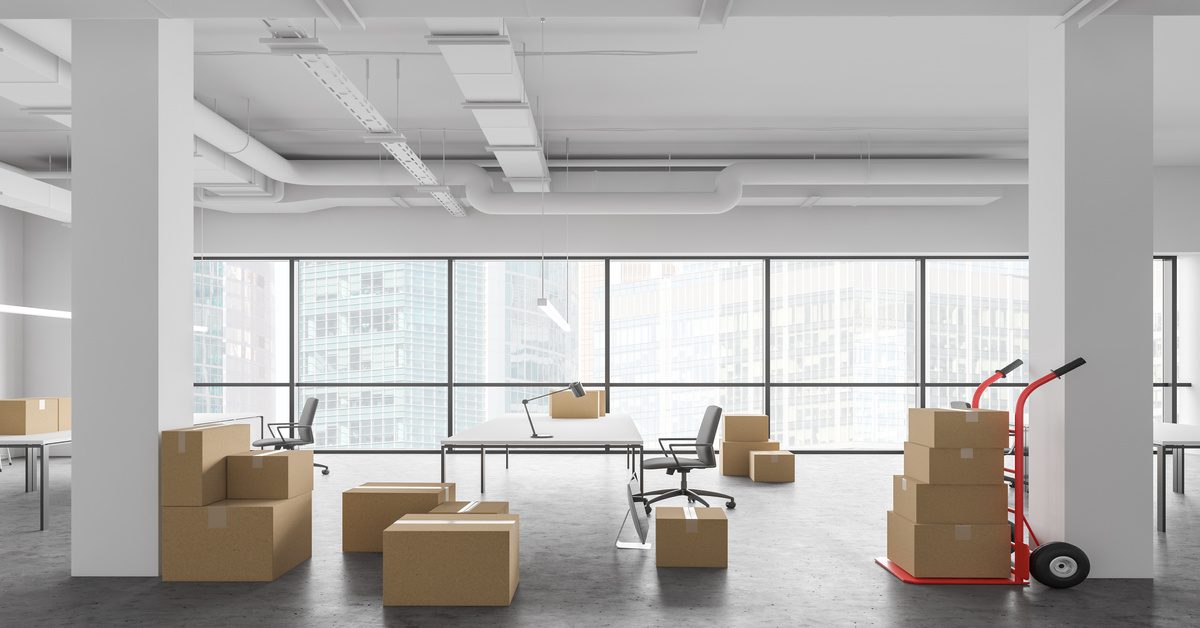 Multiple stacks of cardboard boxes on the gray floor of an open office space with white tables, chairs, walls, and columns.