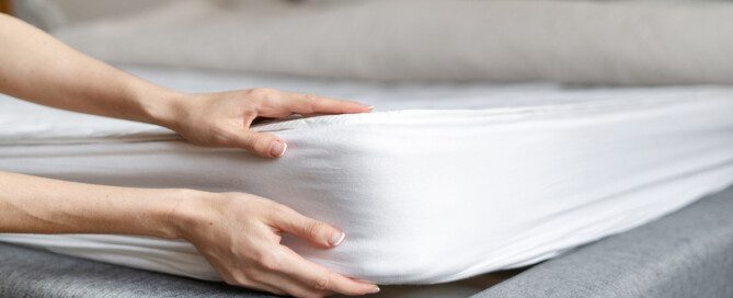 A close-up of an individual lifting a mattress with a white fitted sheet on a gray upholstered bed frame.