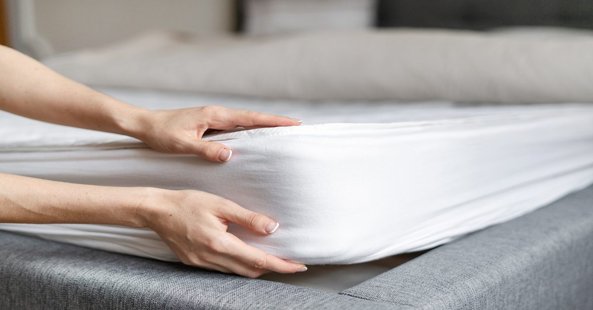 A close-up of an individual lifting a mattress with a white fitted sheet on a gray upholstered bed frame.