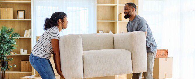 A man and woman smile at each other as they each hold one side of a sofa to move it into their new home.