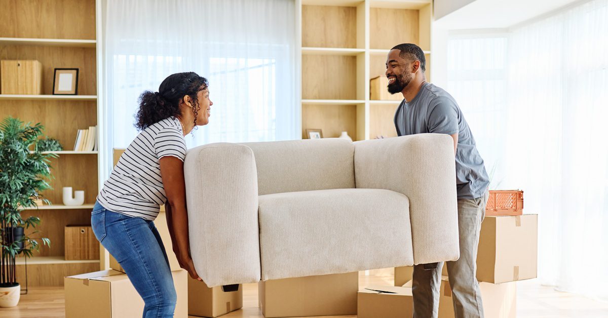 A man and woman smile at each other as they each hold one side of a sofa to move it into their new home.