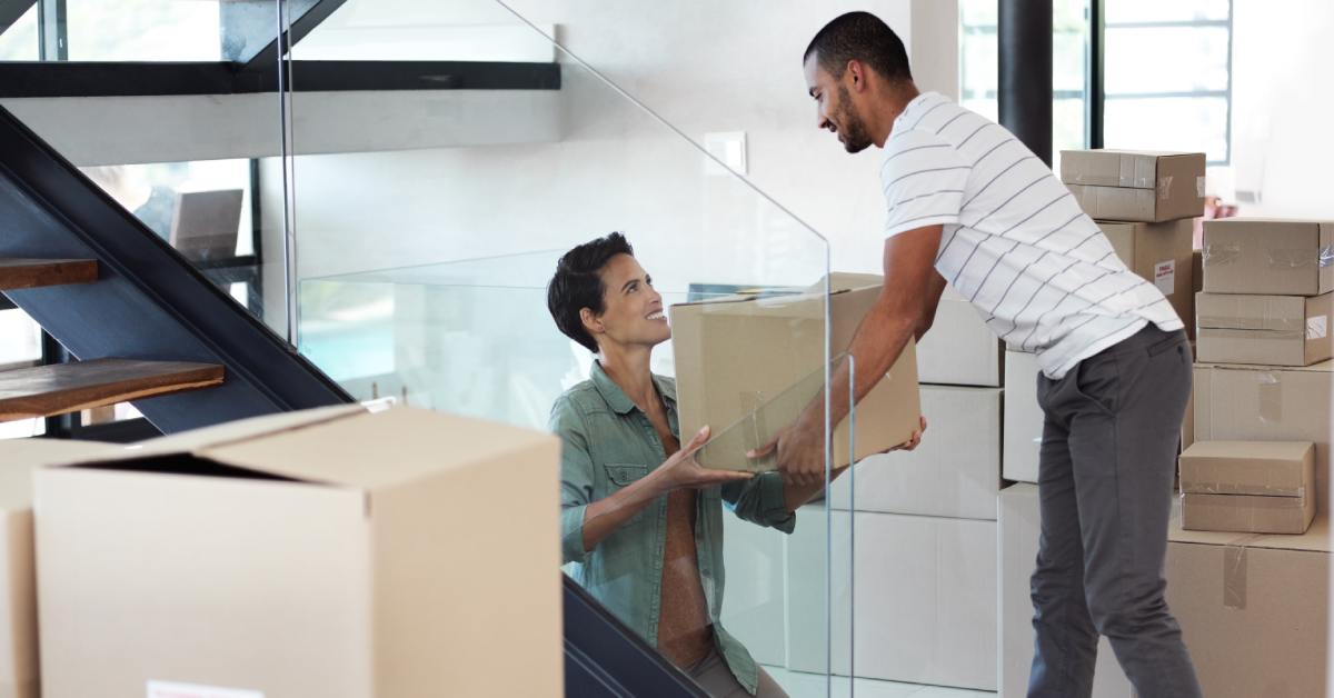 A man and woman exchange a moving box through on modern glass staircase in a bright home surrounded by cardboard boxes.