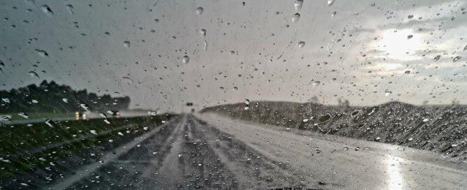 The view from the drivers seat shows rainy windshield and a wet road. The sky is dark and cloud covered in the distance.
