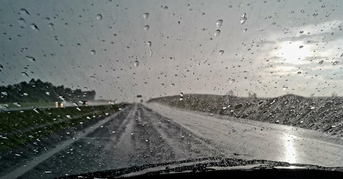 The view from the drivers seat shows rainy windshield and a wet road. The sky is dark and cloud covered in the distance.