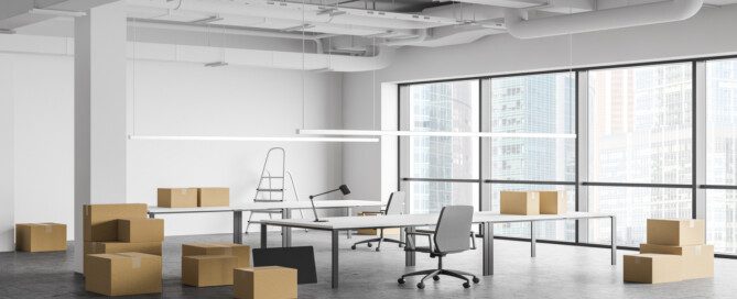 An empty modern office contains large windows, exposed ceilings, desks, chairs, and stacked cardboard boxes.