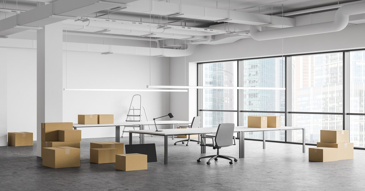 An empty modern office contains large windows, exposed ceilings, desks, chairs, and stacked cardboard boxes.