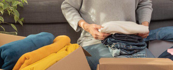 A person sits on the floor packing clothes into two open cardboard boxes, with a sofa and a houseplant in the background.