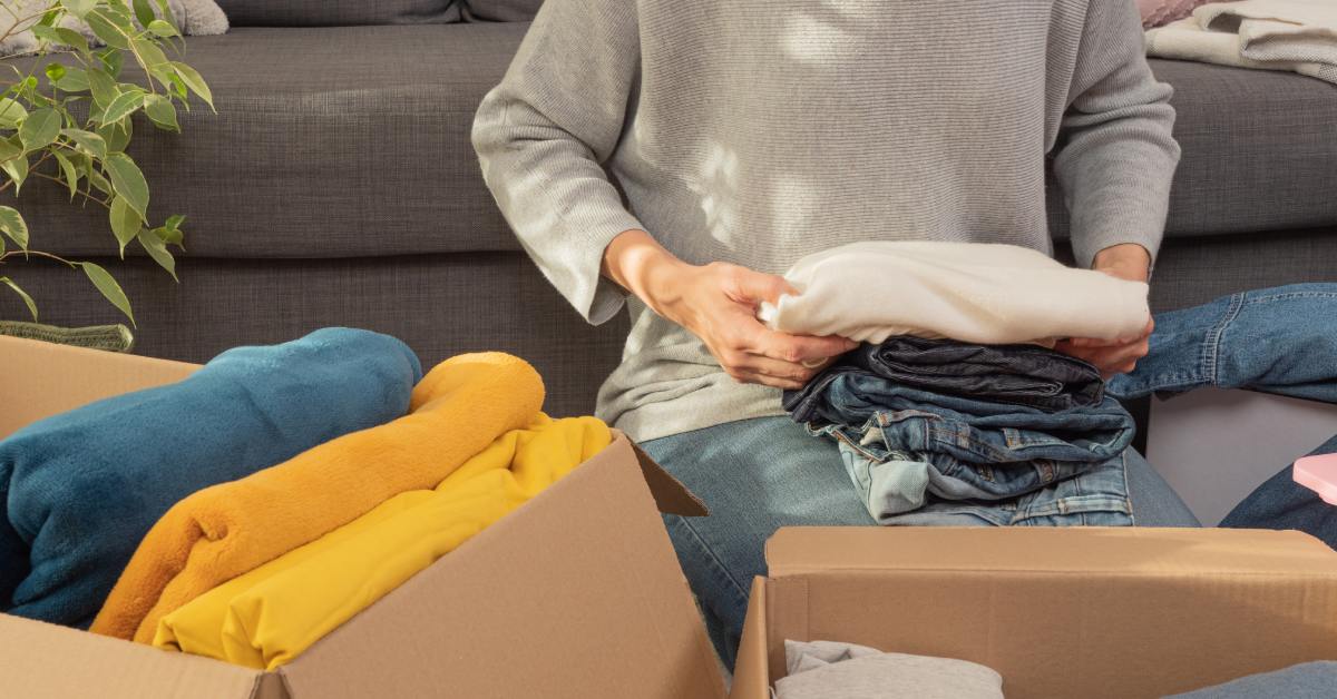 A person sits on the floor packing clothes into two open cardboard boxes, with a sofa and a houseplant in the background.
