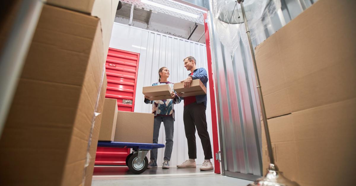 Two smiling people move boxes in a storage unit. A hand truck with stacked boxes and a floor lamp lie nearby.