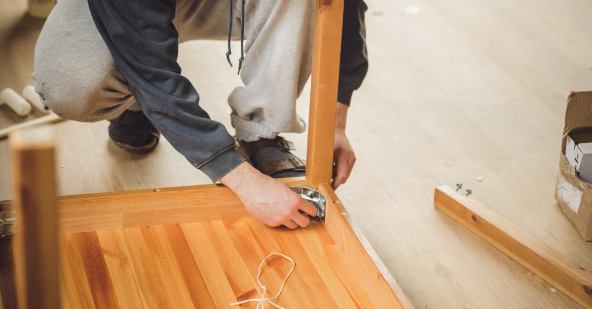 A person disassembles light wood furniture on a wooden floor. Tools and packaging lie scattered nearby.