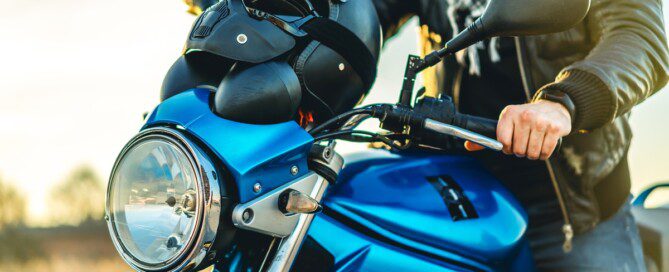 A person wearing a leather jacket sits on a blue motorcycle with a black helmet on the tank while outdoors at sunset.