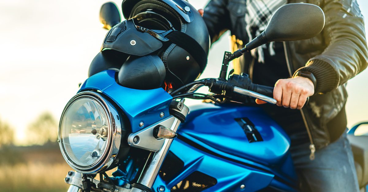 A person wearing a leather jacket sits on a blue motorcycle with a black helmet on the tank while outdoors at sunset.