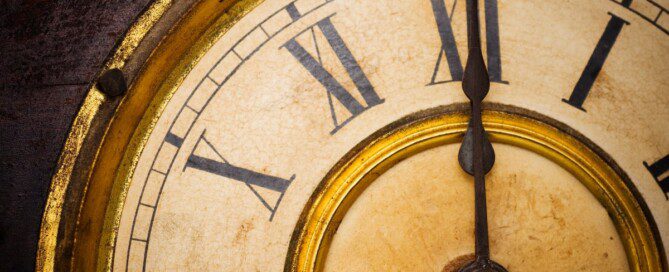 A close-up of an antique clock face featuring black Roman numerals, ornate hands, a cream surface, and tarnished gold bezel.