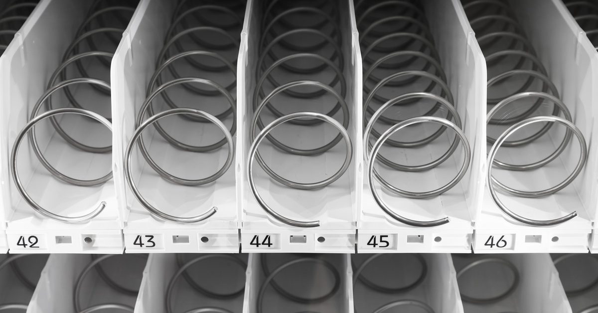 Empty metal spiral coils line numbered rows inside a vending machine, showing white plastic dividers.
