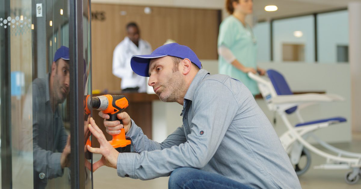 A technician kneels while installing a vending machine with a power drill in a hospital hallway as staff walk behind him.