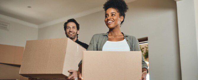A smiling couple carries large cardboard moving boxes into a bright living room in their new apartment.