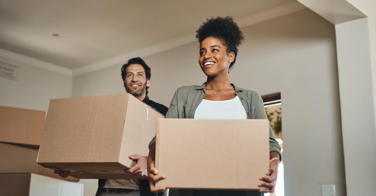 A smiling couple carries large cardboard moving boxes into a bright living room in their new apartment.