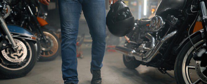 A man wearing jeans and holding a helmet walks through his garage while several large motorcycles sit nearby.