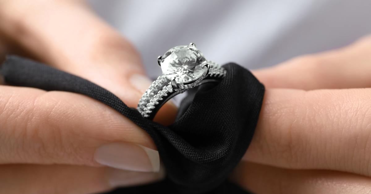 Two hands hold a shiny diamond engagement ring while polishing the band and gemstone with a black cloth.