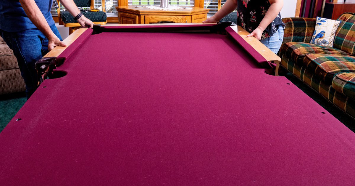 Two people add the rails to a red felt pool table sitting a living room with sofas and wood furniture.