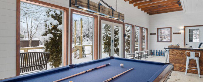 A blue pool table with balls and cues on it sits in a bright room with large windows showing a snowy yard.