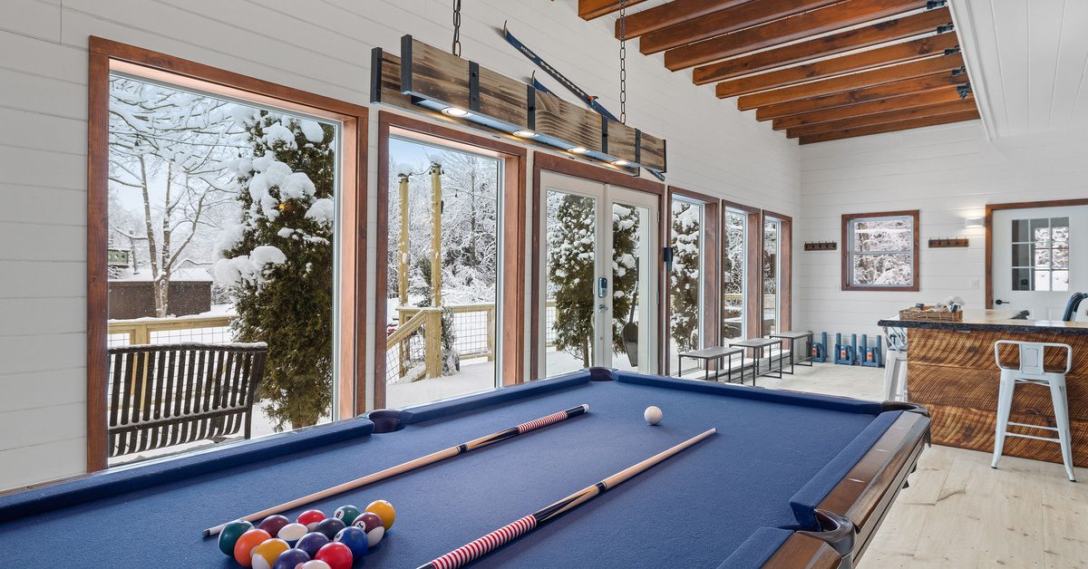 A blue pool table with balls and cues on it sits in a bright room with large windows showing a snowy yard.