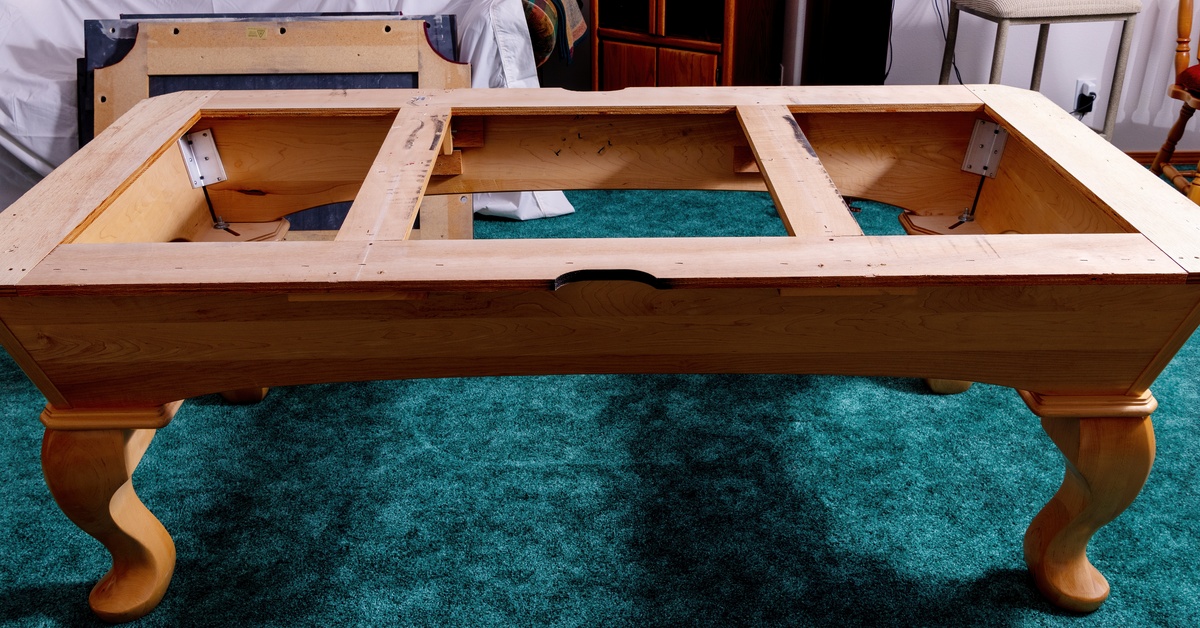 A wooden pool table frame sits disassembled on a green carpet, showing its exposed supports and legs.