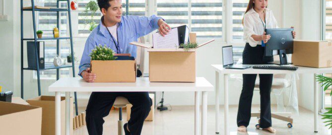 A man packs office supplies into a box at a desk and a woman moves a computer in a bright office during a relocation.