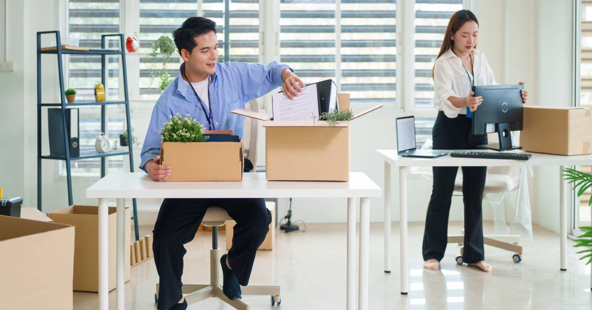 A man packs office supplies into a box at a desk and a woman moves a computer in a bright office during a relocation.