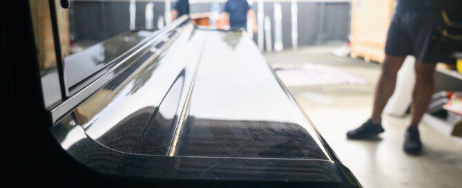 A glossy black piano sits in a storage unit while a mover stands nearby and another person is in the background.