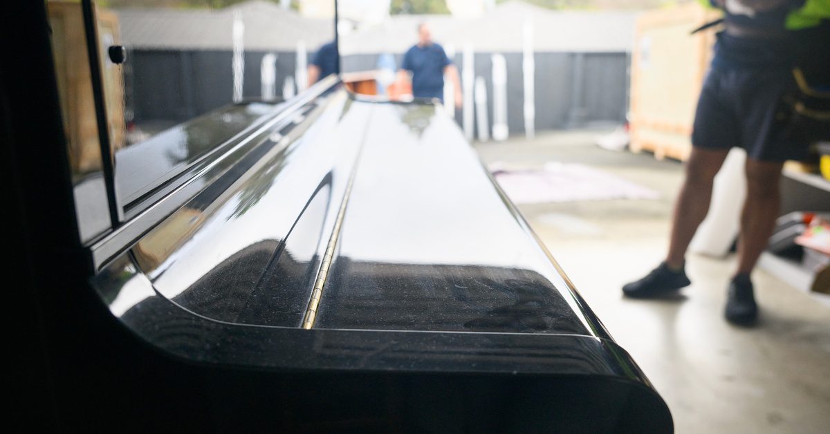 A glossy black piano sits in a storage unit while a mover stands nearby and another person is in the background.