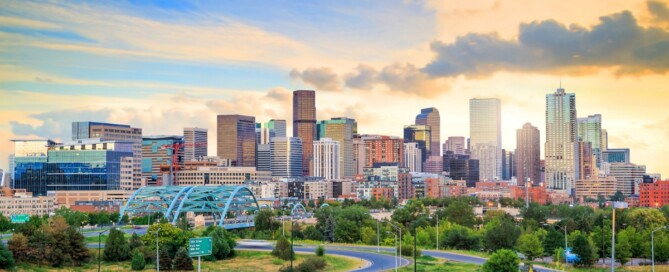 The Denver skyline contains a blue bridge and a curving highway at sunrise, with green trees in the foreground.