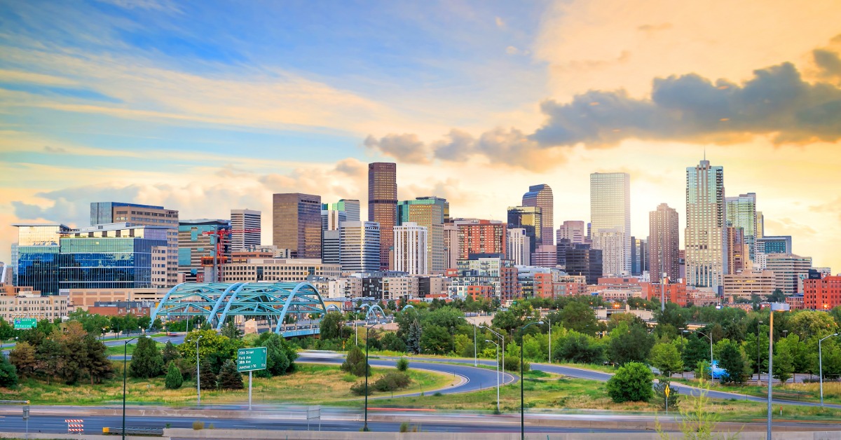 The Denver skyline contains a blue bridge and a curving highway at sunrise, with green trees in the foreground.