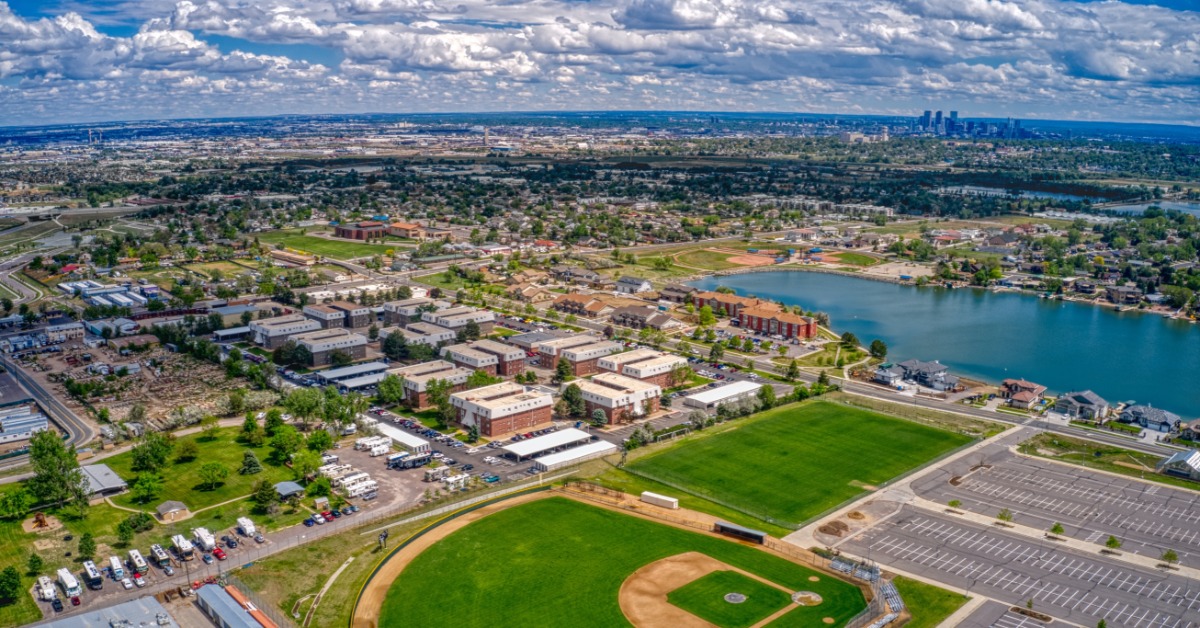An aerial view shows a suburban neighborhood with a baseball field and a lake under a cloudy blue sky.