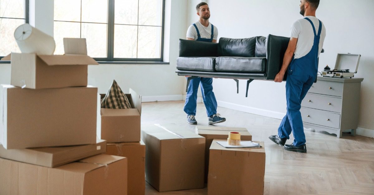 Two movers carry a black sofa across a bright room with stacked boxes, a dresser, and large windows in the background.