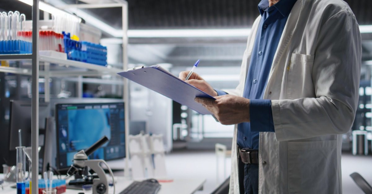 A scientist reviews notes on a clipboard inside a laboratory filled with testing equipment and sample tubes.