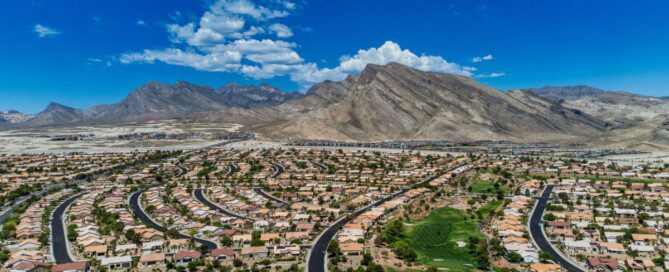 A Las Vegas neighborhood spreads below rugged desert mountains under a bright blue sky with scattered clouds.