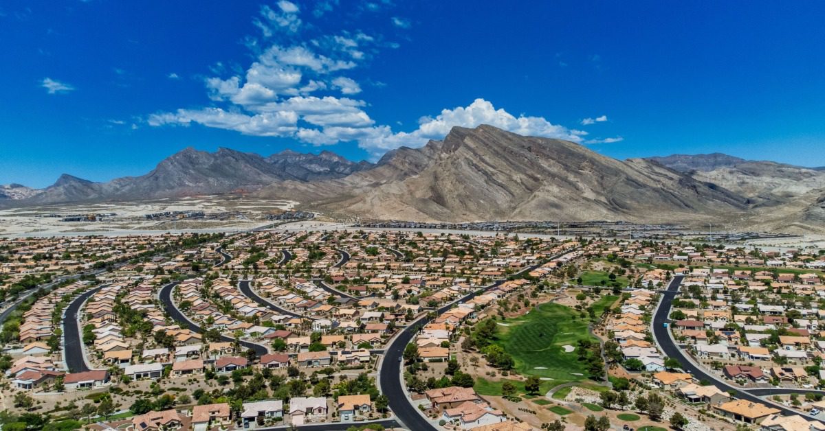 A Las Vegas neighborhood spreads below rugged desert mountains under a bright blue sky with scattered clouds.