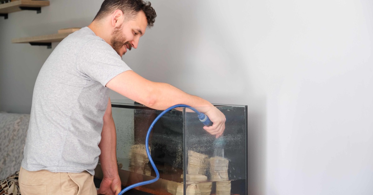 A man wearing a gray shirt and tan pants uses a blue house to pour water into an aquarium with rocks.