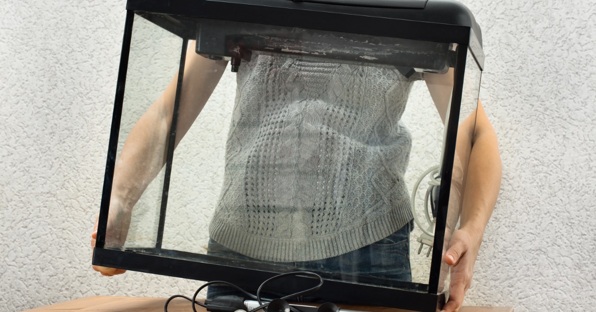 A person carries an empty glass aquarium with cords and fishkeeping tools spread across a wooden table nearby.